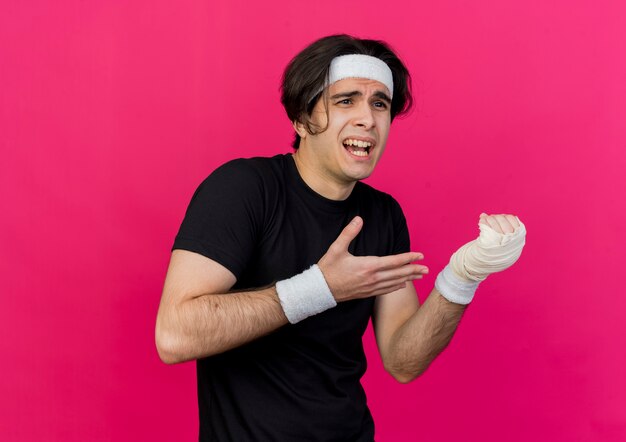 Joven deportivo vistiendo ropa deportiva y diadema apuntando con el brazo a su muñeca vendada sintiendo dolor