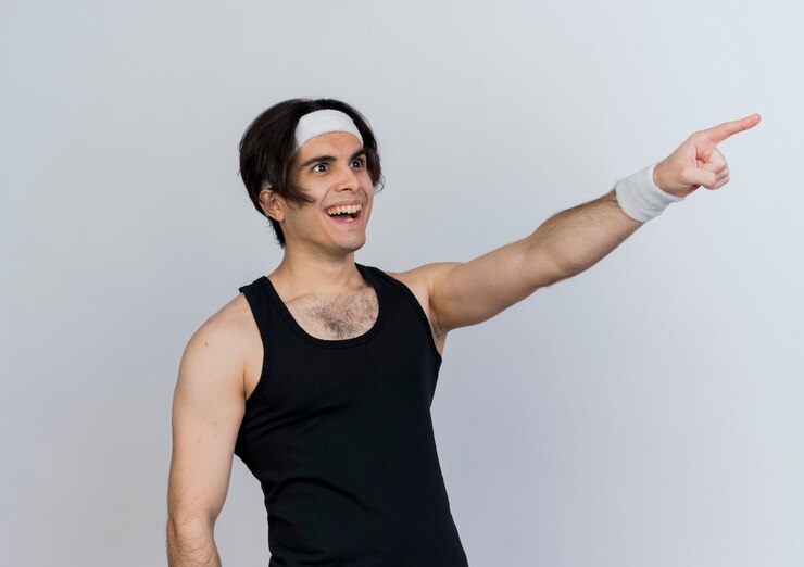 Foto gratuito joven deportivo vistiendo ropa deportiva y diadema apuntando con el dedo índice a algo sonriendo alegremente de pie sobre la pared blanca