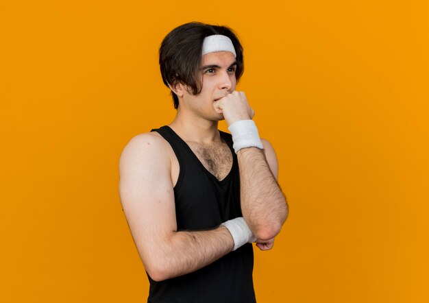 Joven deportivo vistiendo ropa deportiva y diadema con auriculares alrededor del cuello mirando a un lado con la mano en el mentón pensando