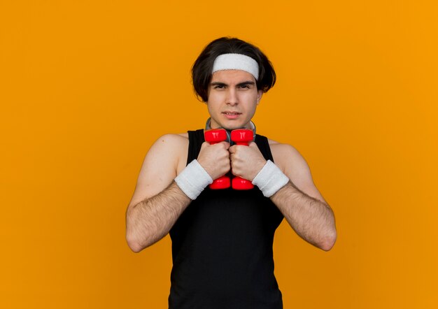 Joven deportivo vistiendo ropa deportiva y diadema con auriculares alrededor del cuello trabajando con pesas mirando confiado con cara seria de pie sobre la pared naranja