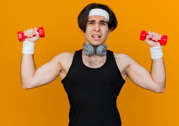 Joven deportivo vistiendo ropa deportiva y diadema con auriculares alrededor del cuello trabajando con pesas mirando confundido