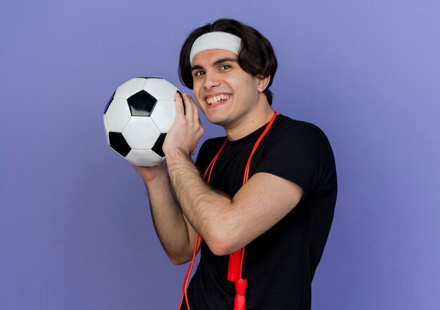Joven deportivo vistiendo ropa deportiva y diadema con cuerda alrededor del cuello sosteniendo un balón de fútbol sonriendo con cara feliz