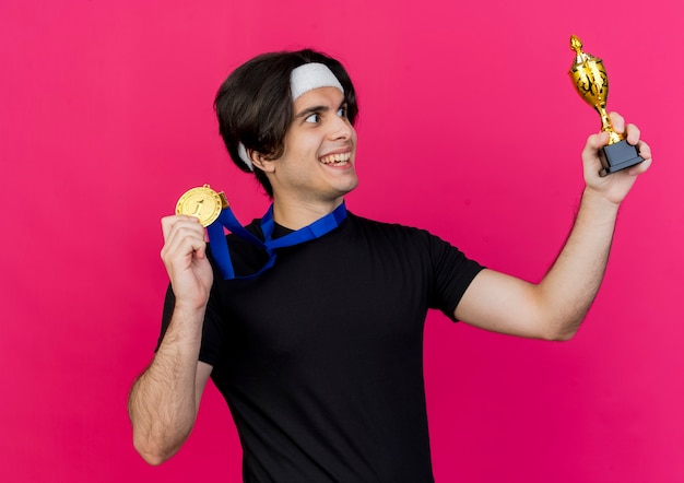 Joven deportivo vistiendo ropa deportiva y diadema con medalla de oro alrededor del cuello mostrando su trofeo feliz y emocionado mirándolo