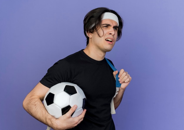 Joven deportivo vistiendo ropa deportiva y diadema con mochila sosteniendo un balón de fútbol lookign a un lado confundido y disgustado