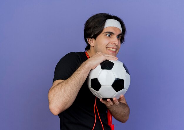 Joven deportivo vistiendo ropa deportiva y diadema con saltar la cuerda alrededor del cuello sosteniendo un balón de fútbol mirando a un lado sonriendo astutamente