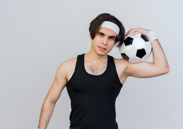 Joven deportivo vistiendo ropa deportiva y diadema sosteniendo un balón de fútbol en el hombro mirando al frente con una sonrisa de confianza de pie sobre la pared blanca