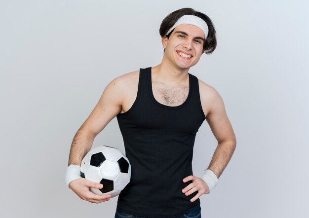 Joven deportivo vistiendo ropa deportiva y diadema sosteniendo un balón de fútbol mirando al frente sonriendo con cara feliz de pie sobre la pared blanca