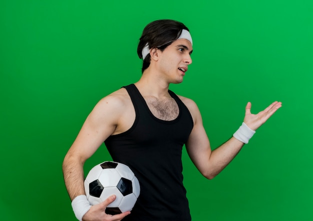 Joven deportivo vistiendo ropa deportiva y diadema sosteniendo un balón de fútbol mirando a un lado con el brazo hacia fuera como pidiendo