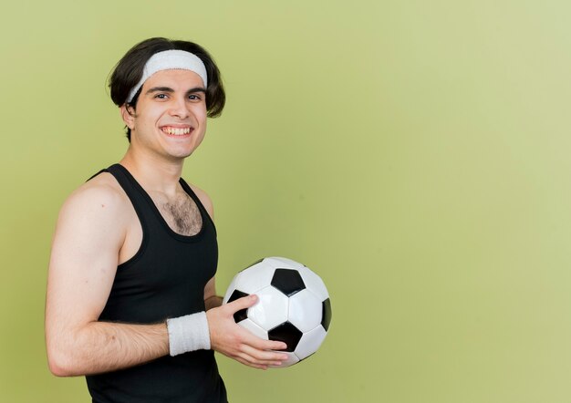 Joven deportivo vistiendo ropa deportiva y diadema sosteniendo un balón de fútbol mirando sonriendo con cara feliz de pie