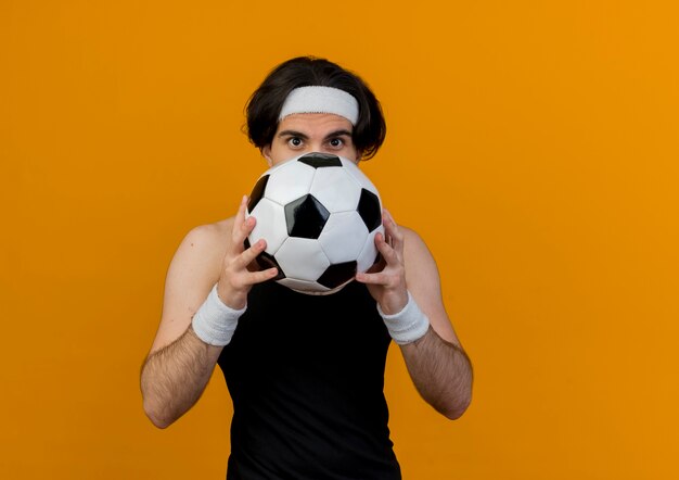 Joven deportivo vistiendo ropa deportiva y diadema sosteniendo un balón de fútbol ocultando su rostro de pie