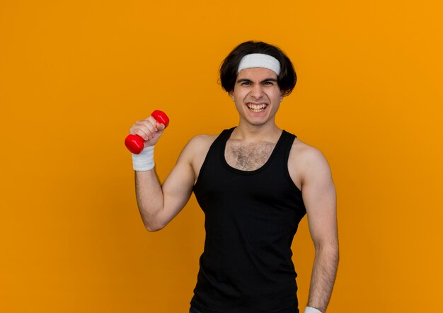 Joven deportivo vistiendo ropa deportiva y diadema sosteniendo mancuernas haciendo ejercicios mirando tenso de pie sobre la pared naranja