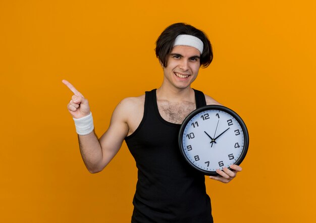 Joven deportivo vistiendo ropa deportiva y diadema sosteniendo un reloj de pared apuntando con el dedo índice hacia el lado sonriendo de pie