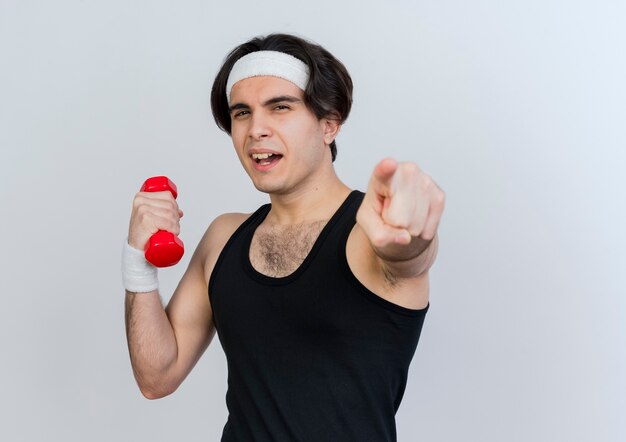 Joven deportivo vistiendo ropa deportiva y diadema trabajando con mancuernas apuntando con el dedo índice al frente sonriendo alegremente de pie sobre la pared blanca