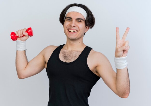 Joven deportivo vistiendo ropa deportiva y diadema trabajando con mancuernas sonriendo con cara feliz mostrando signo v de pie sobre la pared blanca