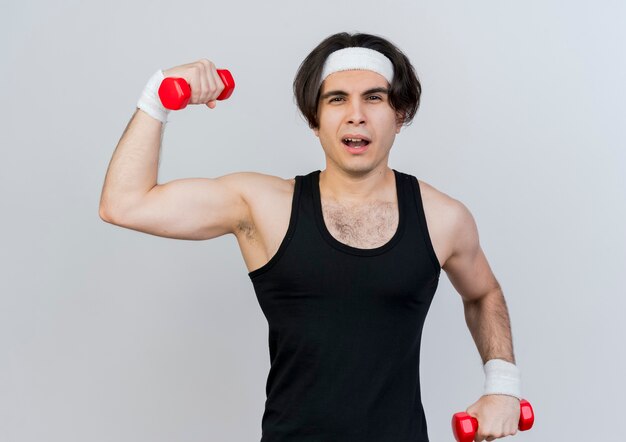 Foto gratuita joven deportivo vistiendo ropa deportiva y diadema trabajando con pesas mirando confiado de pie sobre la pared blanca