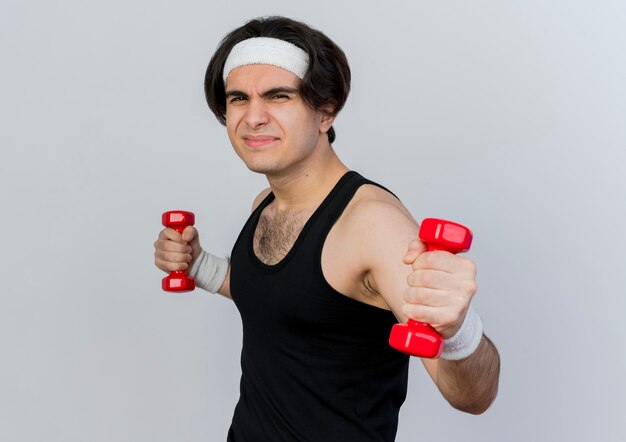 Joven deportivo vistiendo ropa deportiva y diadema trabajando con pesas mirando disgustado de pie sobre la pared blanca