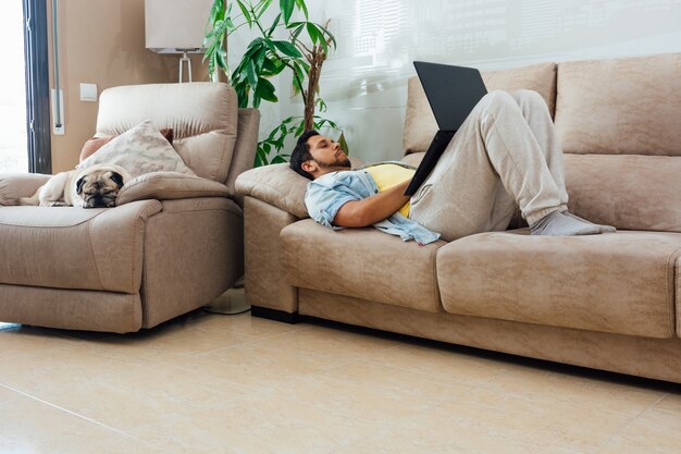 Joven descansando en un sofá en casa y usando una computadora portátil con su perro a su lado