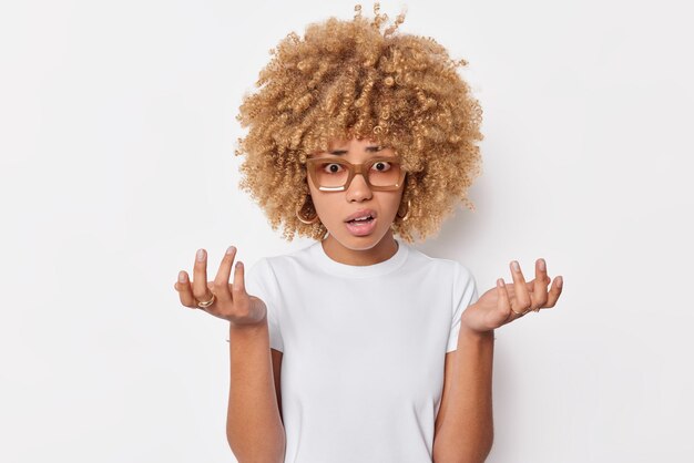 Una joven desconcertada y vacilante con el pelo rizado y tupido extiende las palmas de las manos se siente confundida, se ve avergonzada, usa gafas y una camiseta casual aislada sobre fondo blanco. Entonces que hacer ahora me pregunto