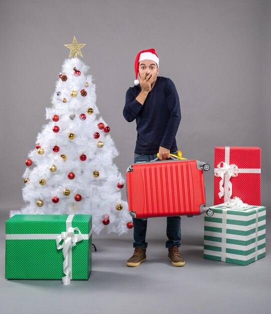 Joven desconcertado con gorro de Papá Noel cerca del árbol de Navidad en gris