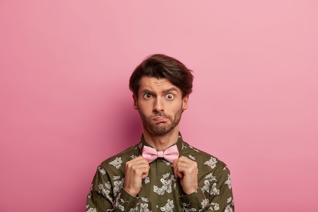 Un joven desconcertado levanta las cejas, tiene cerdas, se ajusta la corbata de moño, usa una camisa de moda con estampado moderno, se viste con buen gusto, se para contra el espacio rosado. Gente