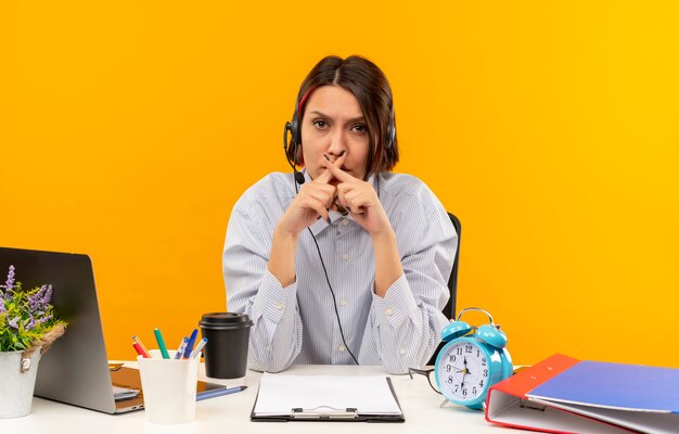 Joven descontento del centro de llamadas con auriculares sentado en un escritorio con herramientas de trabajo sin hacer ningún gesto aislado sobre fondo naranja