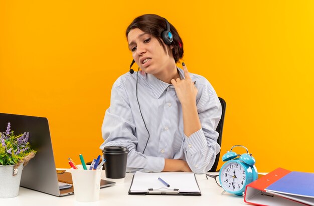 Joven descontento del centro de llamadas con auriculares sentado en un escritorio con herramientas de trabajo levantando el dedo mirando hacia abajo aislado sobre fondo naranja