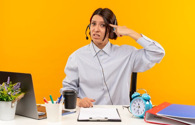 Joven descontento del centro de llamadas con auriculares sentado en el escritorio con herramientas de trabajo poniendo el dedo en la sien aislado sobre fondo naranja