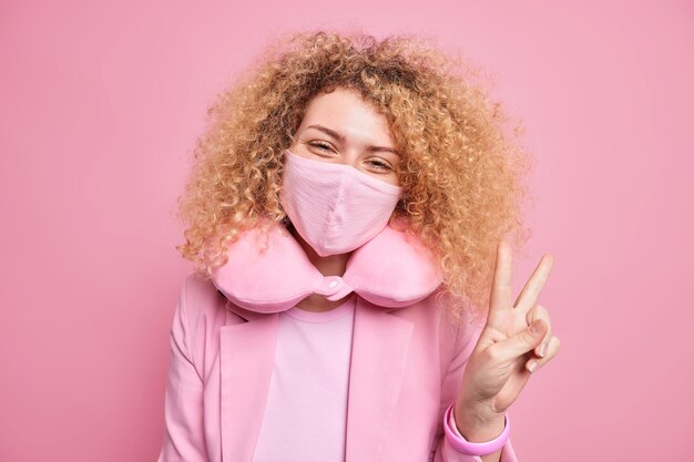 Una joven despreocupada y complacida usa una mascarilla para protegerse del coronavirus, acostumbrada a las medidas de cuarentena, hace el signo de la paz vestida con ropa elegante y usa una almohada para el cuello para sentirse cómodo.