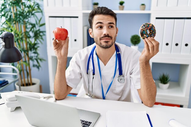 Joven dietista hispano sosteniendo donut y manzana sonriendo mirando hacia un lado y mirando hacia otro lado pensando.