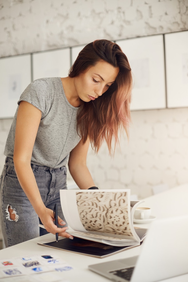 Foto gratuito joven diseñador gráfico hojeando una revista en busca de inspiración para un cliente que usa una computadora portátil.