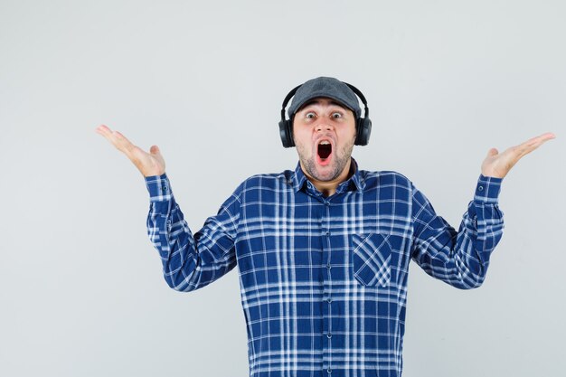 Joven disfrutando de la música con auriculares en camisa, gorra y mirando asombrado. vista frontal.