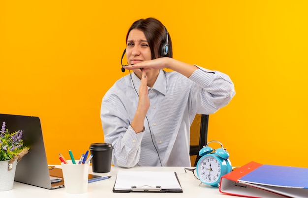 Joven disgustada chica del centro de llamadas con auriculares sentado en el escritorio con herramientas de trabajo haciendo gesto de tiempo de espera aislado sobre fondo naranja