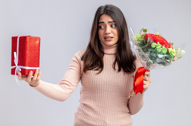 Joven disgustada en el día de San Valentín sosteniendo un ramo mirando la caja de regalo en su mano aislado sobre fondo blanco.