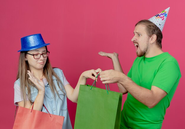 Joven disgustada con sombrero de fiesta azul sostiene una bolsa de regalo roja y levanta la mano tomando la bolsa de regalo verde de un joven molesto con sombrero de fiesta aislado en la pared rosa