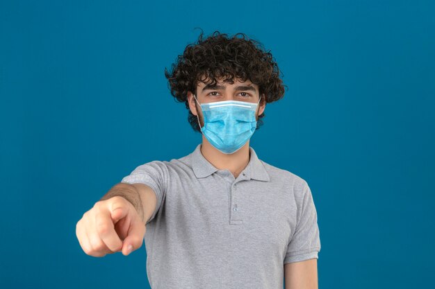 Joven disgustado en máscara protectora médica apuntando a la cámara sobre fondo azul aislado