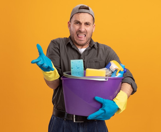 Joven disgustado con ropa casual y gorra en guantes de goma sosteniendo un cubo con herramientas de limpieza levantando la mano con disgusto e indignación de pie sobre la pared naranja