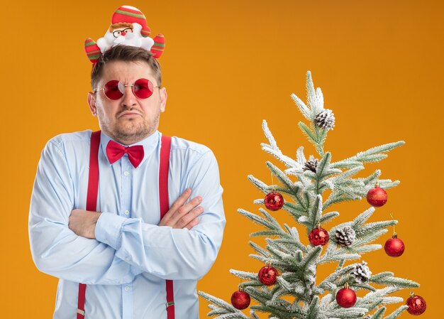 Joven disgustado con tirantes pajarita en el borde con santa y gafas rojas de pie junto al árbol de Navidad mirando a la cámara con los brazos cruzados sobre el pecho sobre fondo naranja