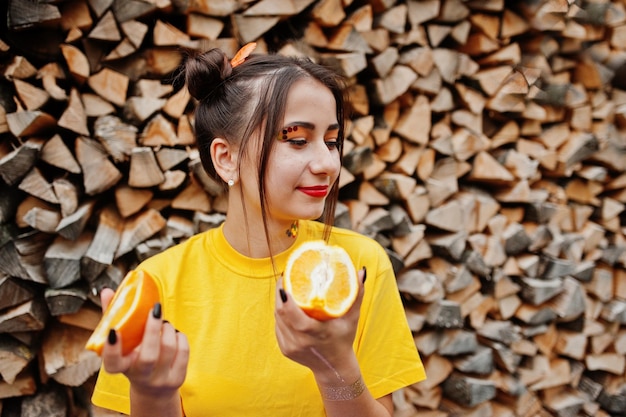 Una joven divertida con un maquillaje brillante en una camisa amarilla sostiene un trozo de naranja sobre un fondo de madera