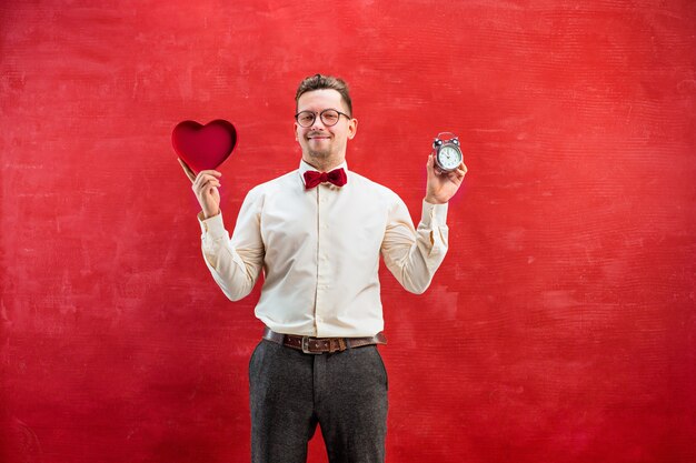 El joven divertido con corazón abstracto y reloj sobre fondo rojo de estudio. concepto - hora de felicitar