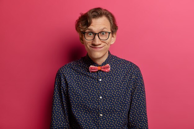 Un joven divertido mira con expresión cómica, usa gafas ópticas y una camisa elegante, nota algo interesante, tiene una conversación agradable con el interlocutor, aislado sobre una pared rosa