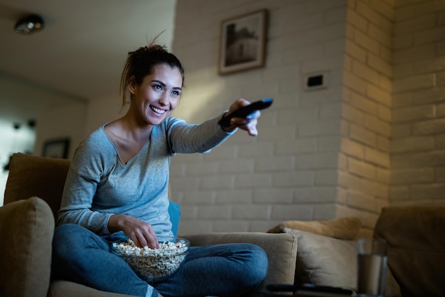 Joven divirtiéndose mientras cambia de canal en la televisión y come palomitas de maíz por la noche en casa