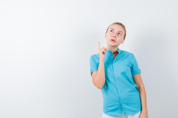 Joven doctora apuntando hacia arriba en uniforme médico, máscara y mirando vacilante