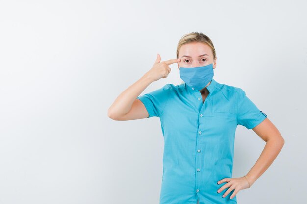 Joven doctora apuntando la cabeza mientras mantiene la mano en la cadera en uniforme aislado