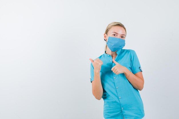 Joven doctora apuntando hacia el lado derecho con los pulgares en uniforme y luciendo bonita