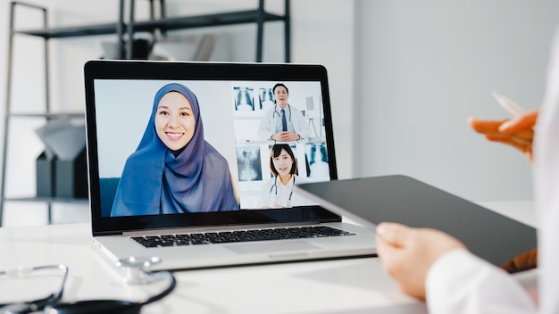 Joven doctora de Asia en uniforme médico blanco con estetoscopio usando computadora portátil hablando por videoconferencia