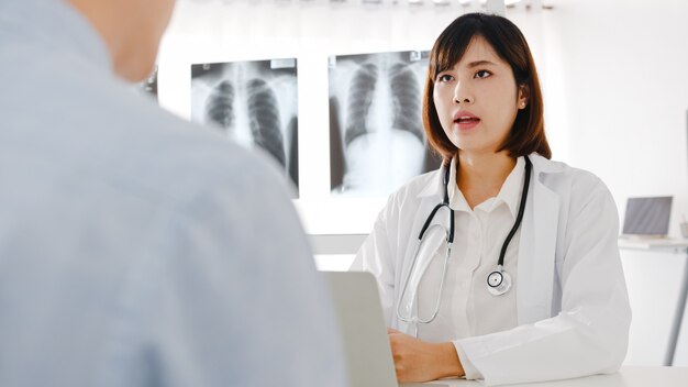 Joven doctora de Asia en uniforme médico blanco usando computadora portátil está brindando excelentes noticias, discuta los resultados