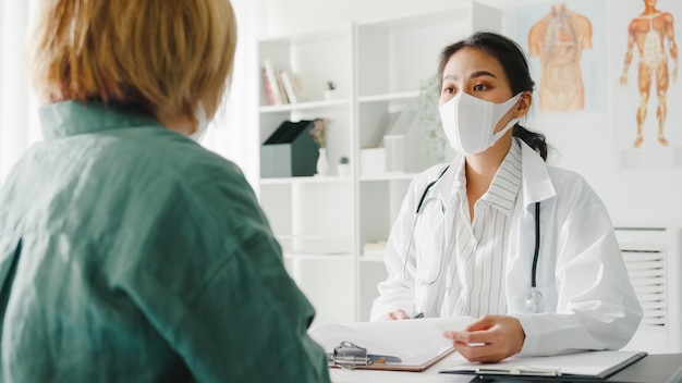 Joven doctora asiática con máscara protectora usando portapapeles discutiendo resultados o síntomas con paciente niña en la oficina del hospital.