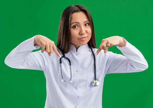 Joven doctora asiática vistiendo bata médica y un estetoscopio mirando a la cámara apuntando hacia abajo en el espacio frente a ella aislado en la pared verde