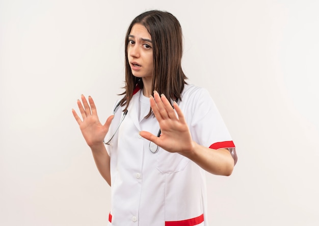 Foto gratuita joven doctora en bata blanca con estetoscopio alrededor del cuello haciendo un gesto de defensa, extendiendo las manos para decirle que no se acerque de pie sobre una pared blanca