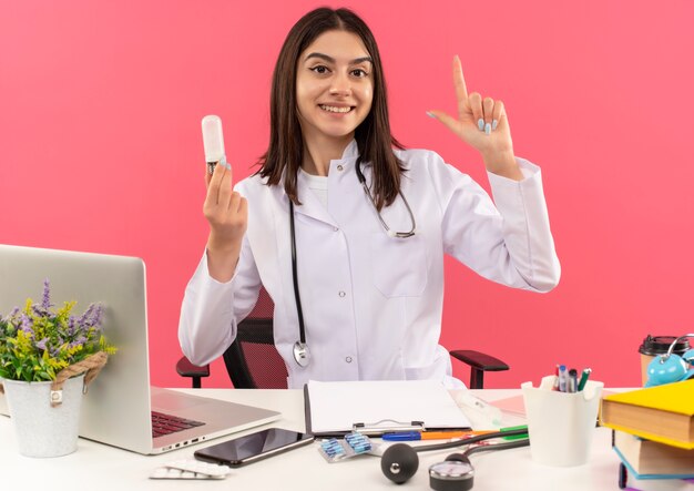 Joven doctora en bata blanca con estetoscopio alrededor del cuello que muestra el bulbo y el dedo índice que tiene una nueva gran idea sonriendo sentado en la mesa con un portátil sobre una pared rosa